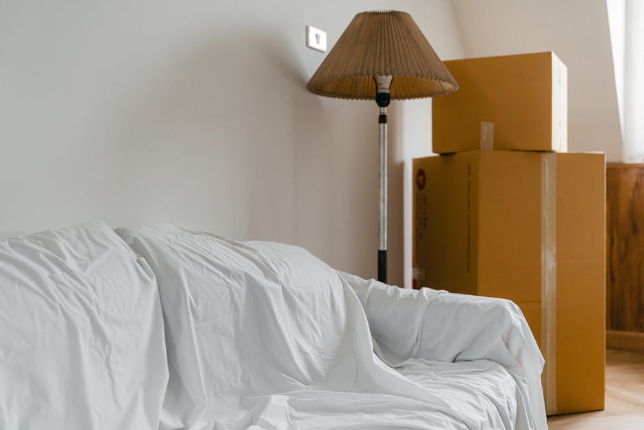 Inside a house preparing for a home relocation, a white sofa draped with a protective cover is positioned near a light grey wall. Closest to the viewer, the sofa's armrest and seat are visible, with the fabric cover slightly wrinkled. In the background, three cardboard boxes of varying sizes are stacked near a wooden cabinet, ready for packing or transport. To the left of the boxes, a tall floor lamp with a brown pleated lampshade is present, casting soft light in the room. The boxes are sealed with packing tape, indicating they are prepared for moving. The scene appears to capture a moment during the packing and moving process, with furniture and belongings organized for transport. Man and Van North Sheen, specialists in removals, often assist in this type of furniture transport and house relocation, ensuring smooth and professional logistical support.