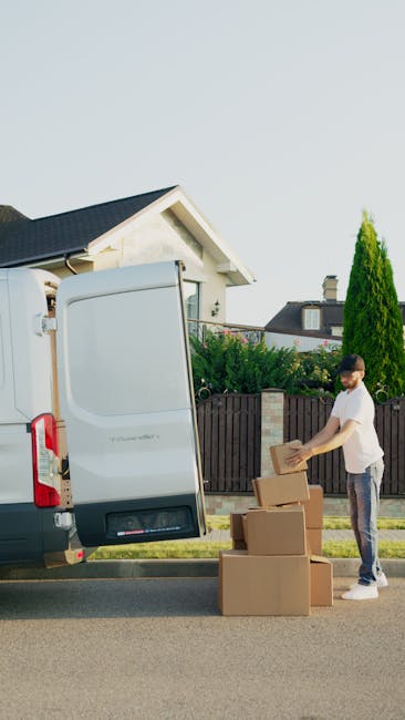 A person wearing a black jacket, dark pants, white sneakers, and a baseball cap is loading or unloading a variety of cardboard boxes into the back of a white moving van parked outdoors on a paved surf