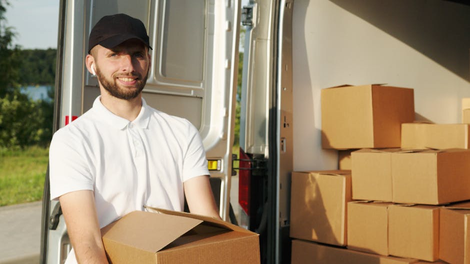 A man with a beard wearing a white polo shirt and a black cap is smiling while holding a cardboard box during a house removal. He is standing outside next to an open van, which is loaded with several stacked cardboard boxes of various sizes, some wrapped in plastic or packing materials. The van is positioned on a paved area with a grassy landscape and trees visible in the background. Metal trolleys and straps are also visible near the loading area, indicating an active loading process for a home relocation. The lighting suggests it is daytime with natural sunlight illuminating the scene. This image illustrates furniture transport and packing activities typical of professional removal services provided by Man and Van North Sheen during a furniture transport or moving operation.