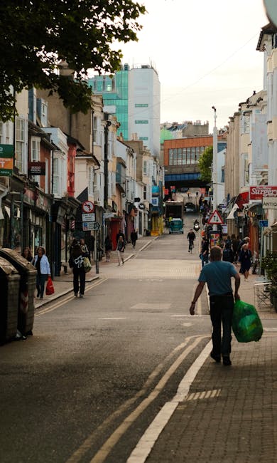 A street scene in a commercial area with a mix of modern and older buildings, including shops with signage and residential stores. The street is paved and slightly inclined, with several pedestrians w