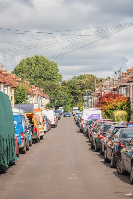 A residential street with lined parked cars on both sides, including small hatchbacks and larger vans, with some covered in protective tarps or blankets as part of a home relocation process. The pavement is clear, and the sky is partly cloudy with a mix of sunlight and shadow. In the background, trees and greenery are visible beyond the row of terraced houses with red brick facades, some with front gardens and bushes. Utility poles and overhead wires run along the street, indicating typical suburban surroundings. This scene depicts the preparation phase of furniture transport and packing during a house removal, with vehicles positioned for loading or unloading, consistent with professional removals services from Man and Van North Sheen.
