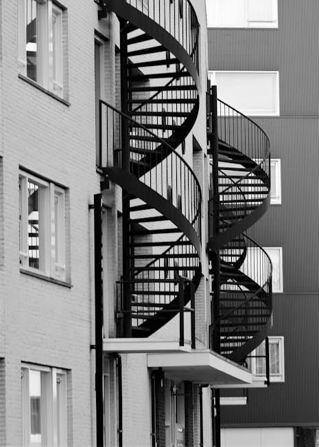 A black metal spiral staircase mounted on the exterior of a multi-storey residential building, featuring open riser steps and handrails on both sides, with nearby windows visible on the building's facade. The staircase connects outdoor landings, providing access between floors for residents or delivery personnel. The surrounding area includes adjacent brick and cladding walls, and the staircase appears to be used in a home relocation or moving process, as part of a professional removals service by Man and Van North Sheen, which specialises in handling challenging access points during house removals. Lighting suggests natural daylight, illuminating the stair structure and the building exterior. The staircase's design allows for efficient furniture and box transport when interior access is restricted.