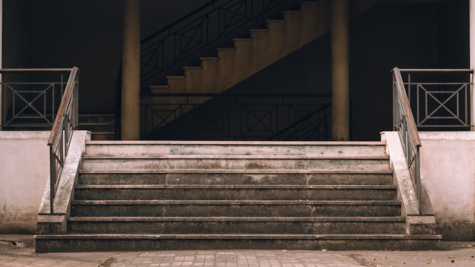 A wide concrete staircase leading up to a building entrance, featuring eight weathered steps with visible dirt and wear, flanked by metal railings on both sides. The staircase is situated outdoors, with a paved ground at the base and some fallen leaves scattered around. Behind the stairs, there are two supporting columns and a dark background showcasing a secondary staircase and railing structure within the building’s interior. The scene is illuminated by natural daylight, highlighting the textures of the concrete and metal. This image reflects typical challenges in house removals where staircase access may impact the logistics of moving furniture and boxes, and is associated with professional removal services like those provided by Man and Van North Sheen, especially concerning staircase access problems and home relocation solutions.
