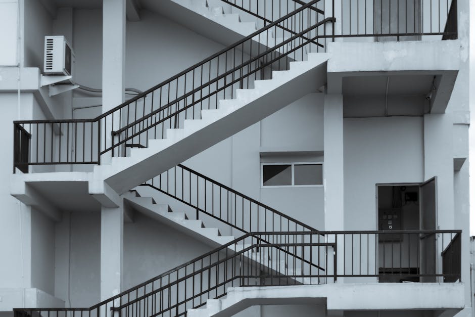 A black metal spiral staircase mounted on the exterior of a multi-storey residential building, featuring open riser steps and handrails on both sides, with nearby windows visible on the building's facade. The staircase connects outdoor landings, providing access between floors for residents or delivery personnel. The surrounding area includes adjacent brick and cladding walls, and the staircase appears to be used in a home relocation or moving process, as part of a professional removals service by Man and Van North Sheen, which specialises in handling challenging access points during house removals. Lighting suggests natural daylight, illuminating the stair structure and the building exterior. The staircase's design allows for efficient furniture and box transport when interior access is restricted.