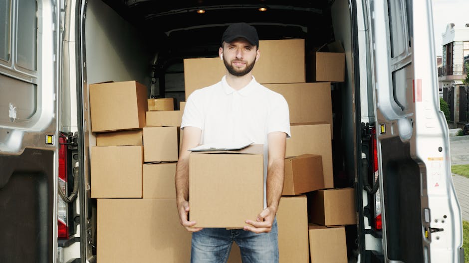 A man with a beard and wearing a black cap, white t-shirt, and jeans is standing inside the open rear of a large white van, which is parked on a paved street near residential buildings. He is holding a medium-sized cardboard box, with several other boxes of various sizes stacked around him inside the van. The boxes are sealed with packing tape and appear to be prepared for a home relocation. The van's interior is configured for furniture transport and packing, with some boxes stacked near the back doors and others arranged along the sides. Outside, the environment suggests daylight, and the scene depicts a loading process typical of house removals, with additional objects like protective packing materials likely present but not visible in the frame. The image relays a professional moving and packing service, exemplified by Man and Van North Sheen, emphasising their home removals and furniture transport activities.