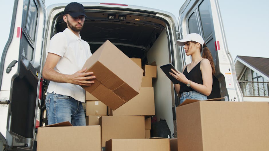 Two individuals are engaged in a home relocation or furniture transport process with a white moving van parked outside a residential property. The man, wearing a white polo shirt, jeans, and a dark cap, is inside the van holding a medium-sized cardboard box, preparing to load or unload it. The woman, dressed in a black sleeveless top, light blue jeans, and a white cap, stands beside the open vehicle door, holding a clipboard or tablet, likely for inventory or packing checks. The interior of the van is filled with neatly stacked cardboard boxes of various sizes, some wrapped with plastic film or packing materials, indicating an active packing or unpacking phase. The surrounding environment includes a paved driveway and a house with a sloped roof and balcony visible in the background, under daylight conditions. This scene depicts the logistics involved in home removals, emphasizing tasks like packing, furniture transport, and loading processes that are typical during house moves, as managed by companies such as Man and Van North Sheen.