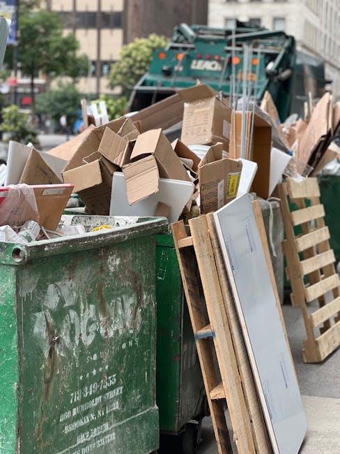 A large green waste disposal bin placed on a city street, filled with various types of packaging materials including flattened cardboard boxes, open on top with some overflowing. Several smaller cardboard boxes and packaging remnants are visible spilling over the edges of the bin. Nearby, a wooden pallet is leaning against the side of the bin, supporting a large flat-screen television wrapped in protective plastic film. In the background, a green waste collection truck is parked, partially obscured by the bin, with trees, modern buildings, and parked vehicles also visible. The scene suggests a disposal area used for clearing packing materials and household waste related to home relocation or furniture transport, which Man and Van North Sheen occasionally assist with as part of their removals services. The environment appears to be outdoors in an urban setting with natural and built elements, and daylight illuminates the scene clearly.