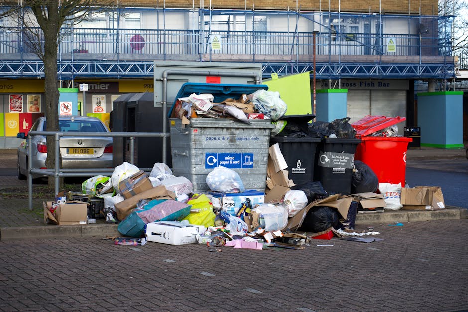 Outside a commercial building in North Sheen, a community waste disposal area with multiple overflowing bins is visible. The scene includes a large grey mixed paper and cardboard recycling container filled with crumpled paper, cardboard boxes, and plastic packaging, with some contents spilling onto the surrounding paved surface. Next to it, there are black, green, and red refuse bins containing various waste types, including plastic bags, paper, and other rubbish. Cardboard boxes, some flattened and others partially assembled, are scattered on the ground nearby. A metal railing separates the waste area from a parked silver car on the left side of the image. In the background, the building has a scaffolding structure, large windows, and signs for nearby shops, including a fish bar. The overall scene depicts a typical waste disposal spot that might be relevant to house removals or relocation services in North Sheen, captured in daylight with natural lighting conditions.